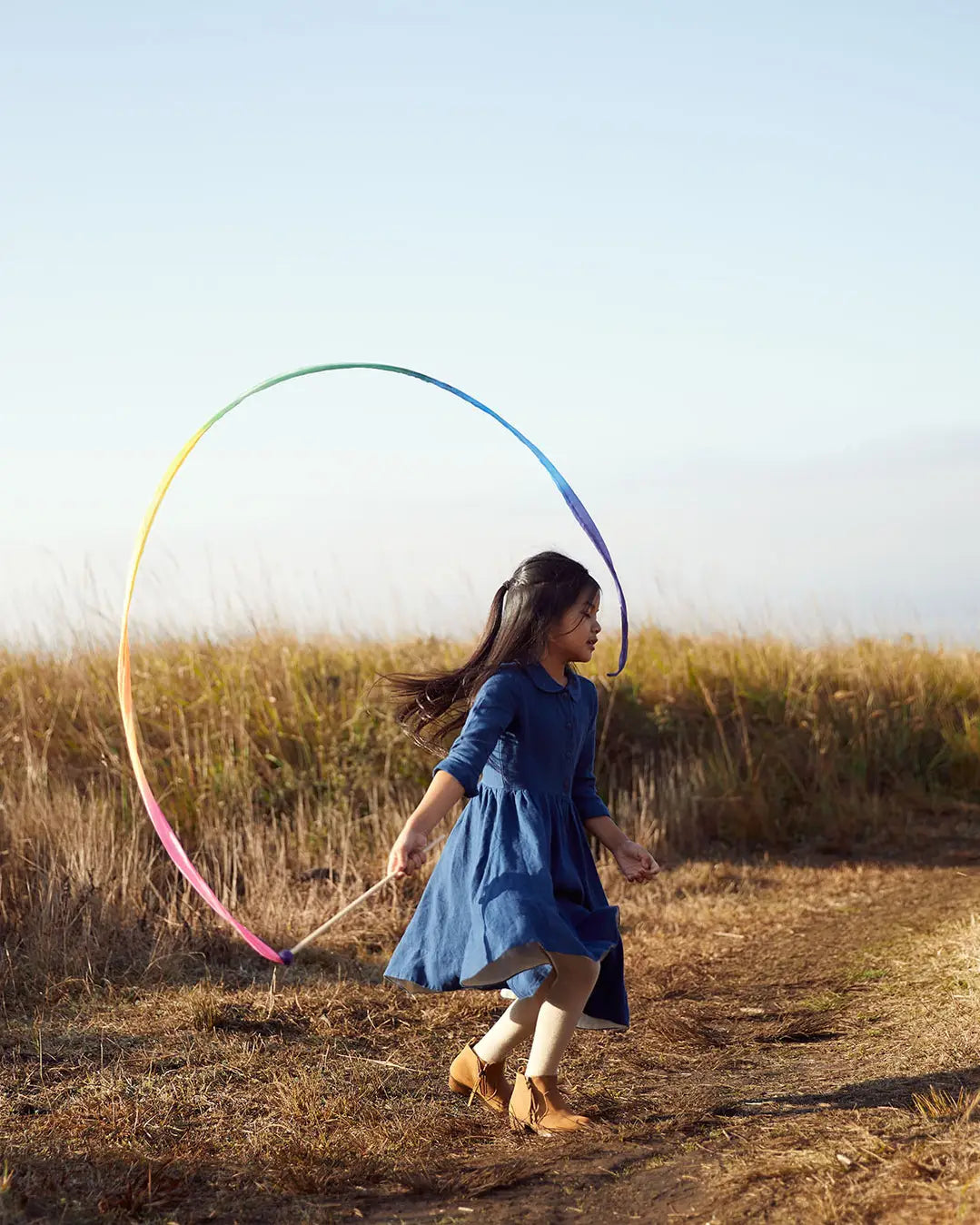 Child running outdoors with a Sarah’s Silks Rainbow Streamer Wand forming a large colorful loop.